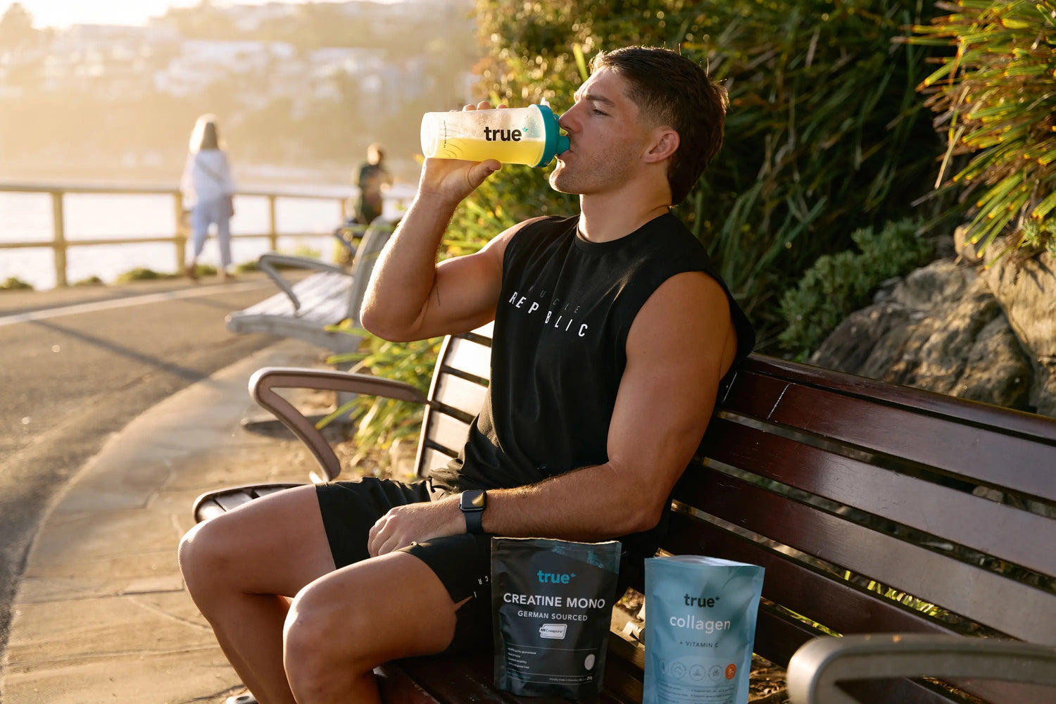 Man drinking from a True Nutrition shaker bottle on a seaside promenade bench at sunset, with True Creatine Mono and True Collagen product pouches beside him — fitness supplement, workout recovery, outdoor coastal workout image.