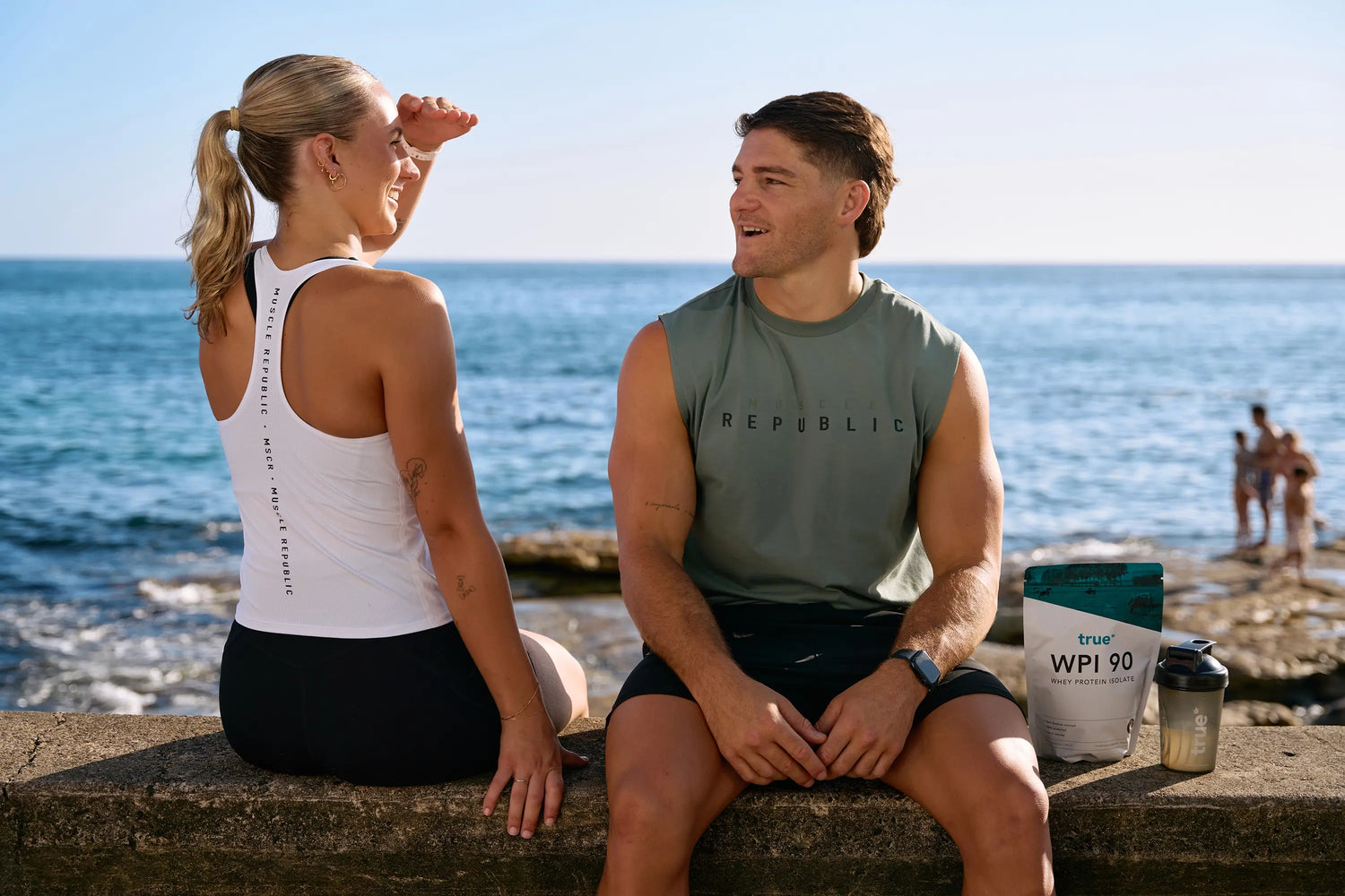 Two fit people in Muscle Republic workout gear sitting on a seaside concrete ledge at sunset, smiling and chatting next to a pouch of True WPI 90 whey protein isolate and a protein shaker—outdoor coastal fitness lifestyle image.