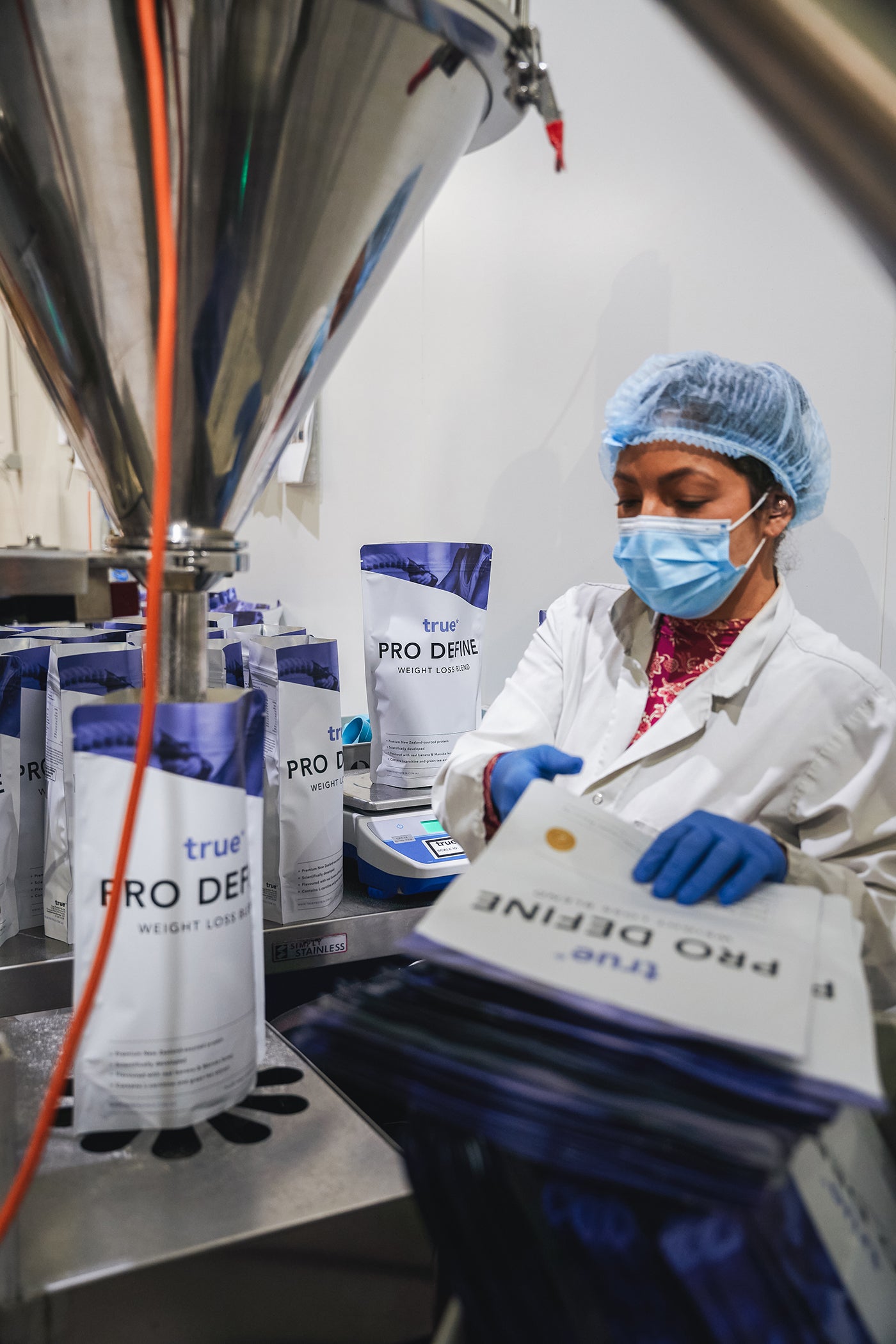 Factory worker in hairnet, mask and gloves packing True Pro Define protein powder weight-loss supplement pouches on a production line in a food manufacturing/packaging facility.