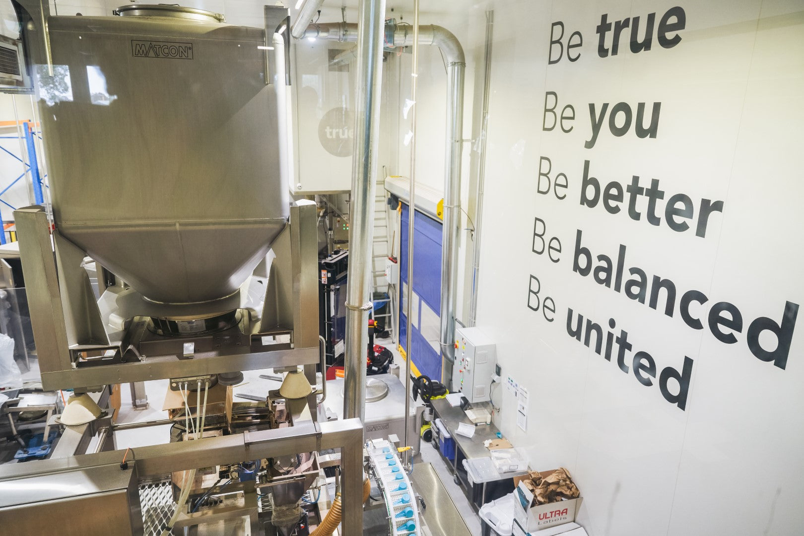 Industrial food manufacturing interior showing stainless-steel hopper and conveyor line in a protein packaging plant with large motivational wall text reading “Be true, Be you, Be better, Be balanced, Be united.”