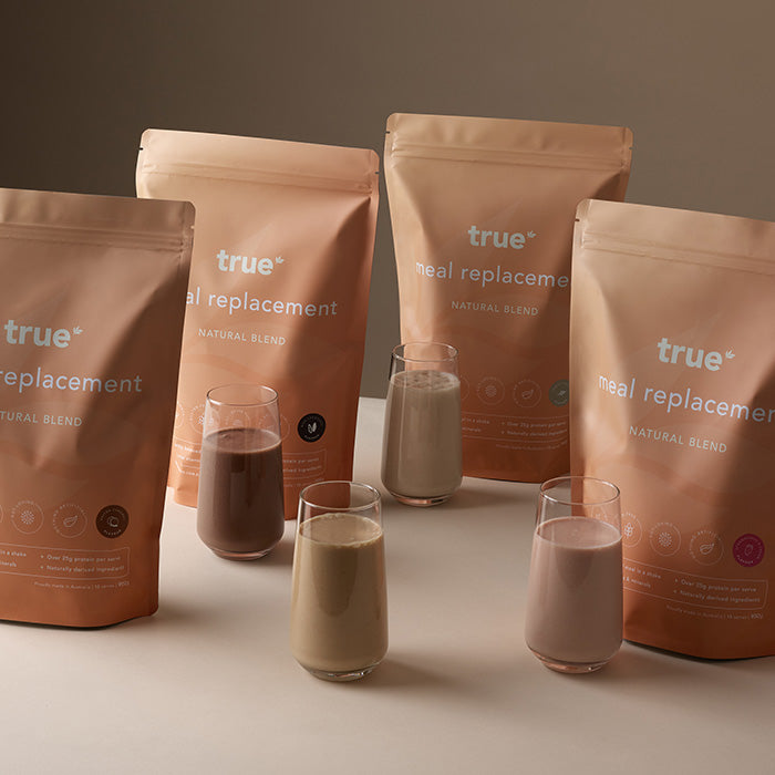 True meal replacement natural blend pouches with five poured protein shake glasses in assorted flavors on a neutral studio background, product packaging and serving display for healthy meal replacement shakes.