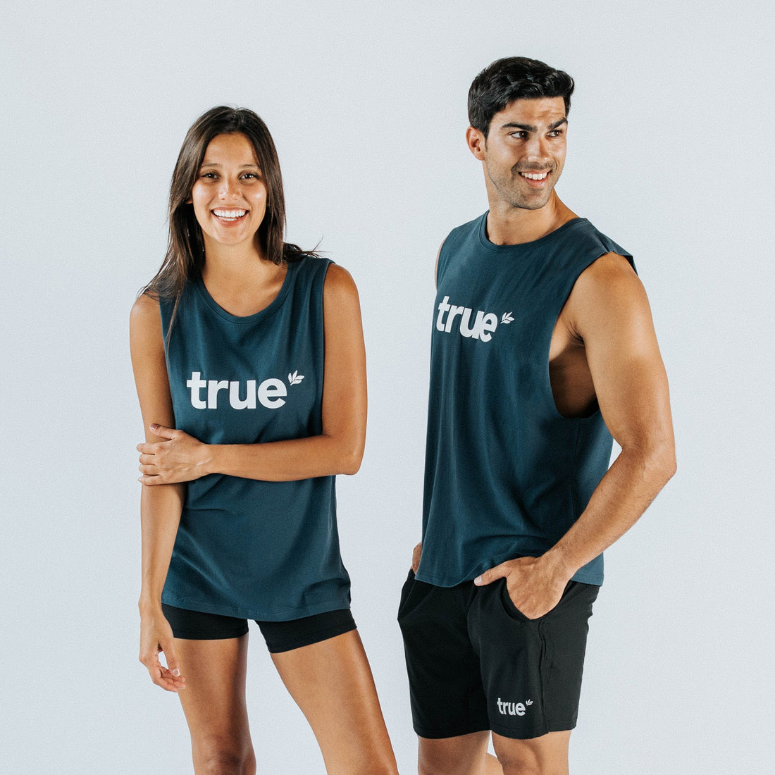 Two models (female and male) wearing navy "true" logo sleeveless athletic tank tops with black shorts — unisex breathable training tanks, studio product photo for online activewear.