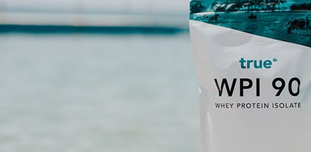 Close-up of True WPI 90 whey protein isolate pouch (protein powder) against a sunlit beach and sea background — premium sports nutrition, whey protein isolate pouch lifestyle image.