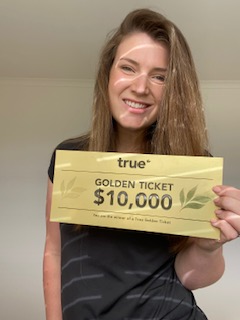 Smiling woman holding a True-branded yellow "Golden Ticket $10,000" giveaway voucher indoors, winner announcement and prize promotion.