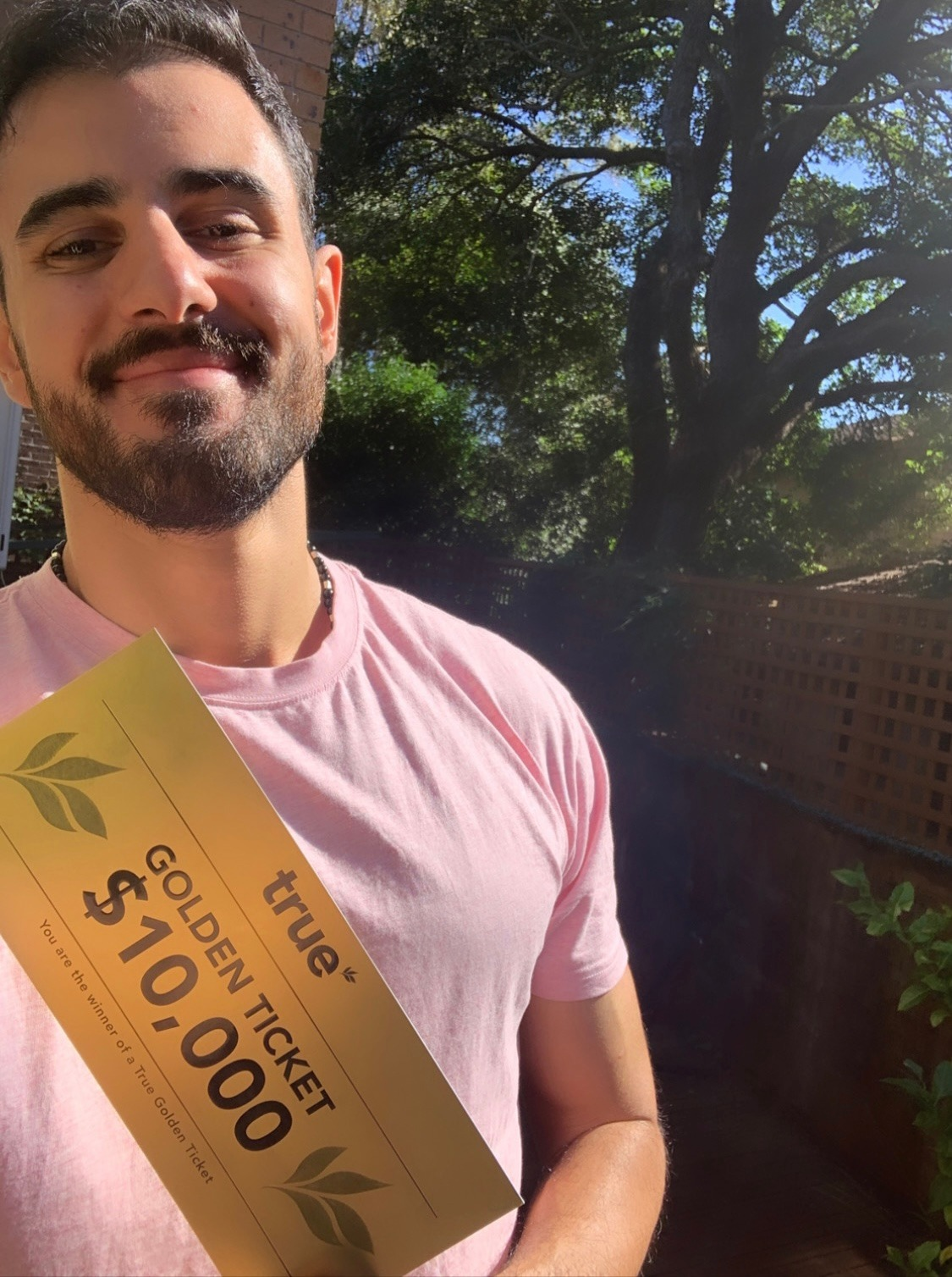 Young man in a pink t-shirt holding a gold "True" $10,000 golden ticket in a sunlit backyard with large oak tree and wooden fence