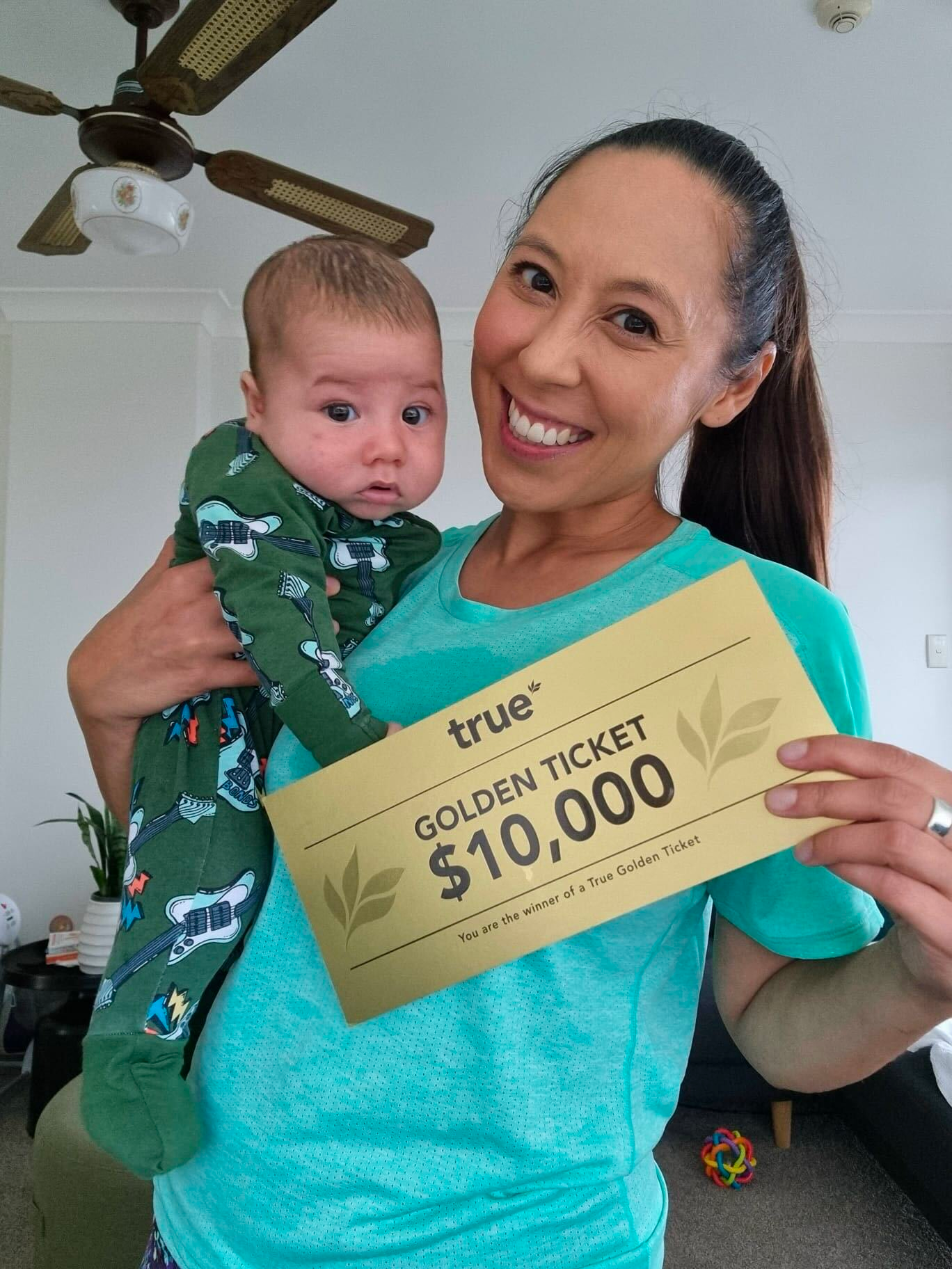 Smiling woman holding baby and gold "true" Golden Ticket reading "$10,000" in a bright living room — happy mother and infant celebrating giveaway prize.