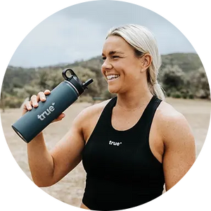 Smiling fit woman in black True tank top holding a teal True insulated stainless-steel water bottle on a rugged outdoor trail/bushland — fitness lifestyle, hiking and reusable water bottle product shot.