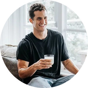 Smiling young man in a black t-shirt sitting on a sofa by a sunlit window, holding a glass of milk in a modern bright living room