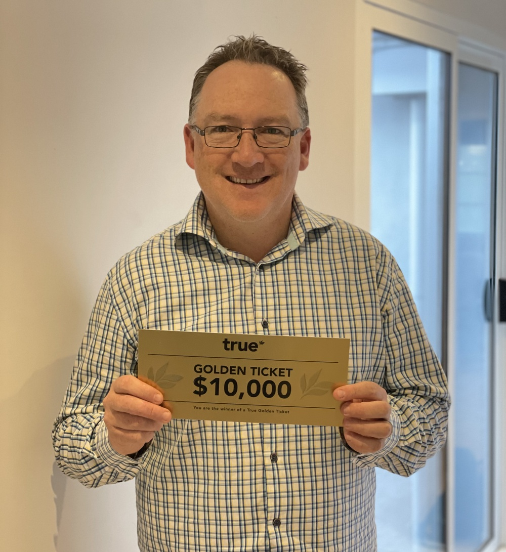 Smiling middle‑aged man wearing glasses and a blue plaid shirt holding a gold “Golden Ticket $10,000” prize voucher inside a bright modern home — winner, cash prize, voucher, promotional portrait.