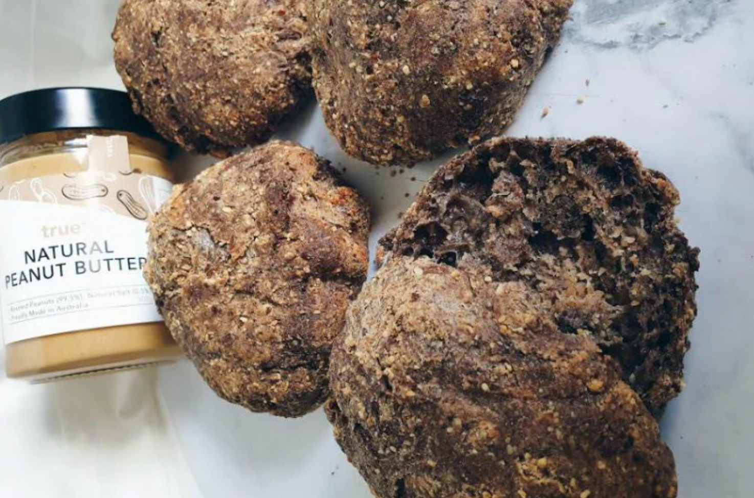 Jar of natural peanut butter next to four wholegrain seeded brown bread rolls on a white marble surface, close-up food product lifestyle shot.