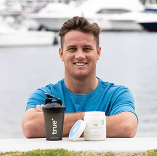 Smiling man in blue shirt at a marina with yachts in background, leaning on a ledge next to a white supplement tub and black "True" shaker bottle — waterfront fitness supplement product photo.