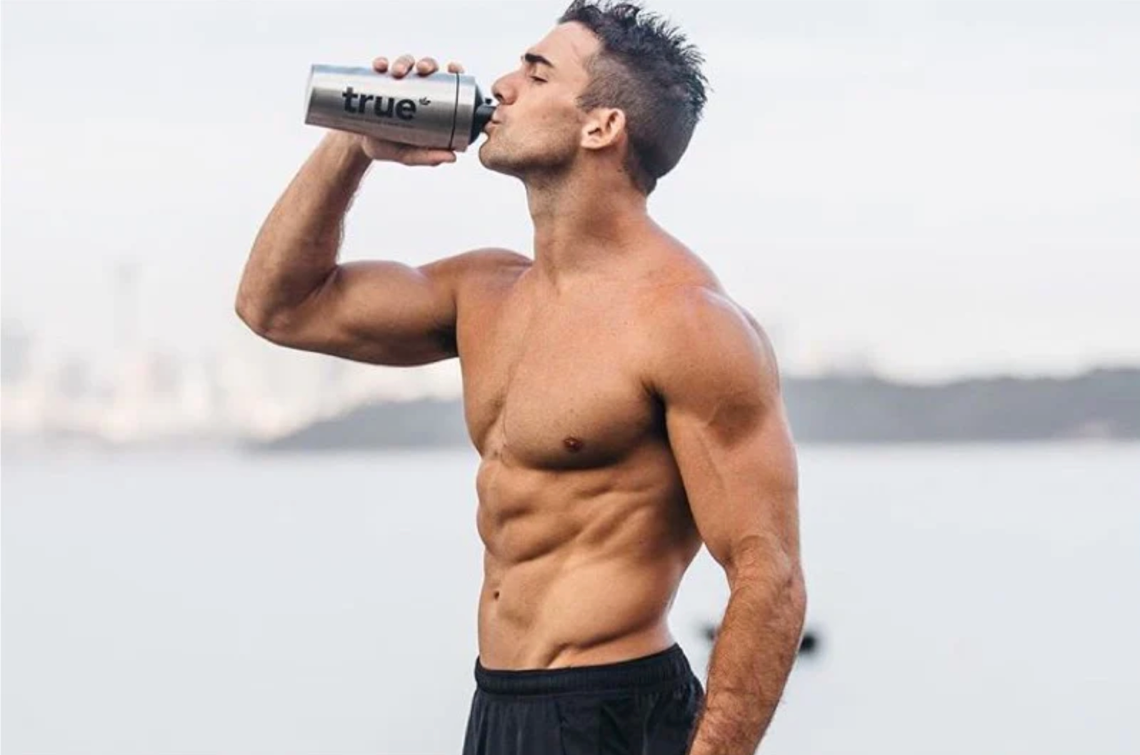 Shirtless fit man drinking from stainless steel "true" shaker bottle at a waterfront beach — muscular abs, outdoor coastal workout and supplement shaker photo.