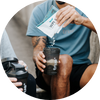 Athlete pouring whey protein powder from a single-serve sachet into a black shaker bottle labeled “true” during an outdoor workout — protein shake, fitness supplement.
